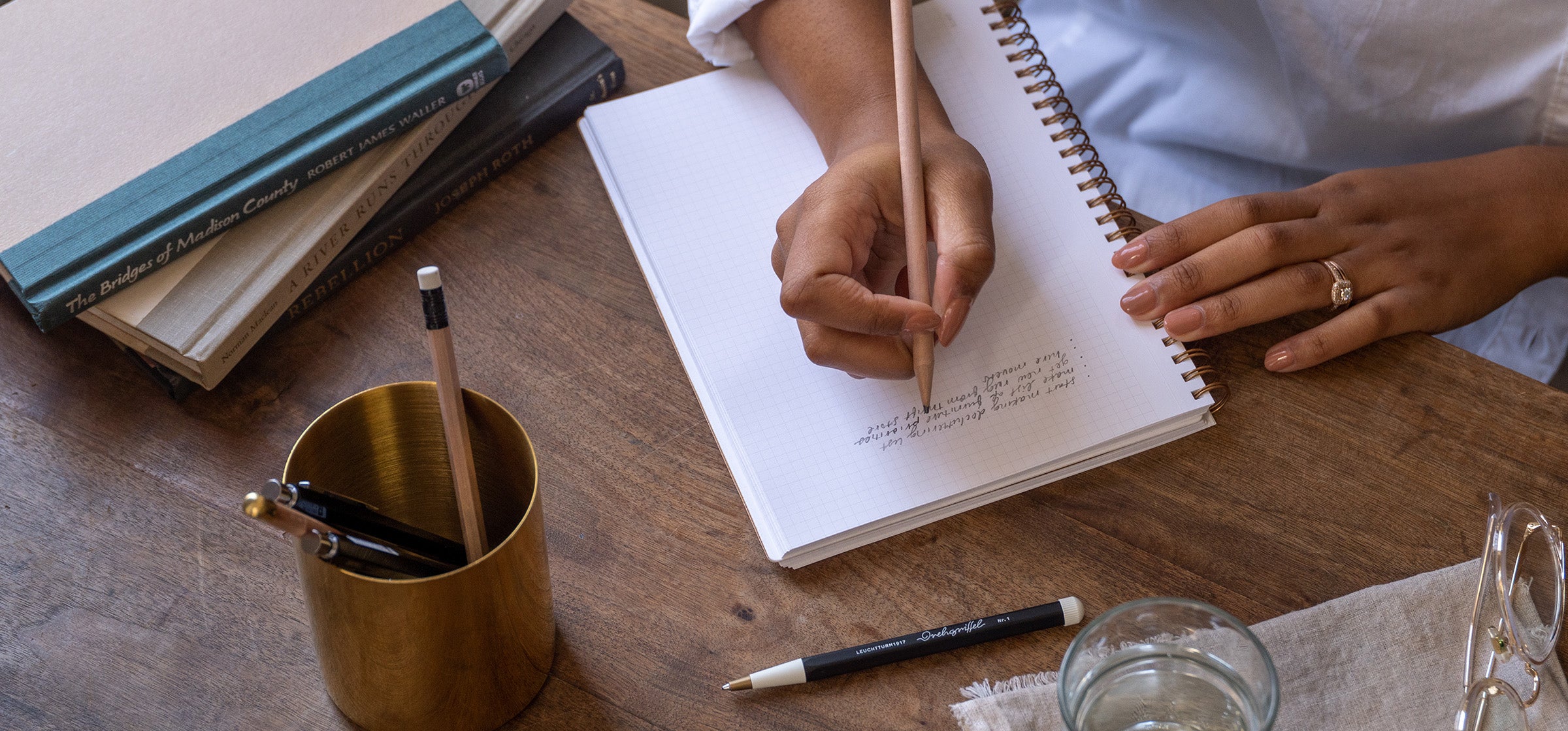 A woman writes in a journal at a cozy wooden desk. Her hand is posed above the paper with a classic no. 2 pencil. A few books, writing tools, and a glass of water are strewn across the desk.