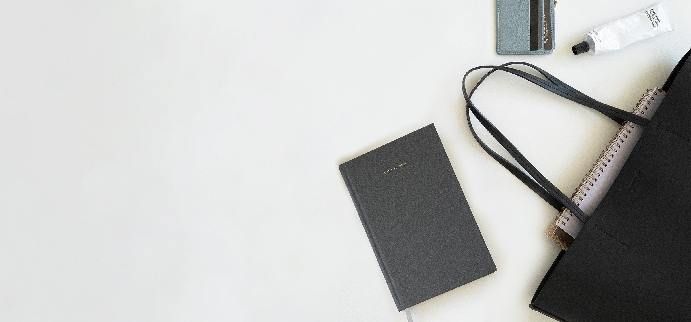 A hardcover daily planner spills out of a black tote bag onto a white surface. Other writing tools, a lip balm, and accessories are also visible.