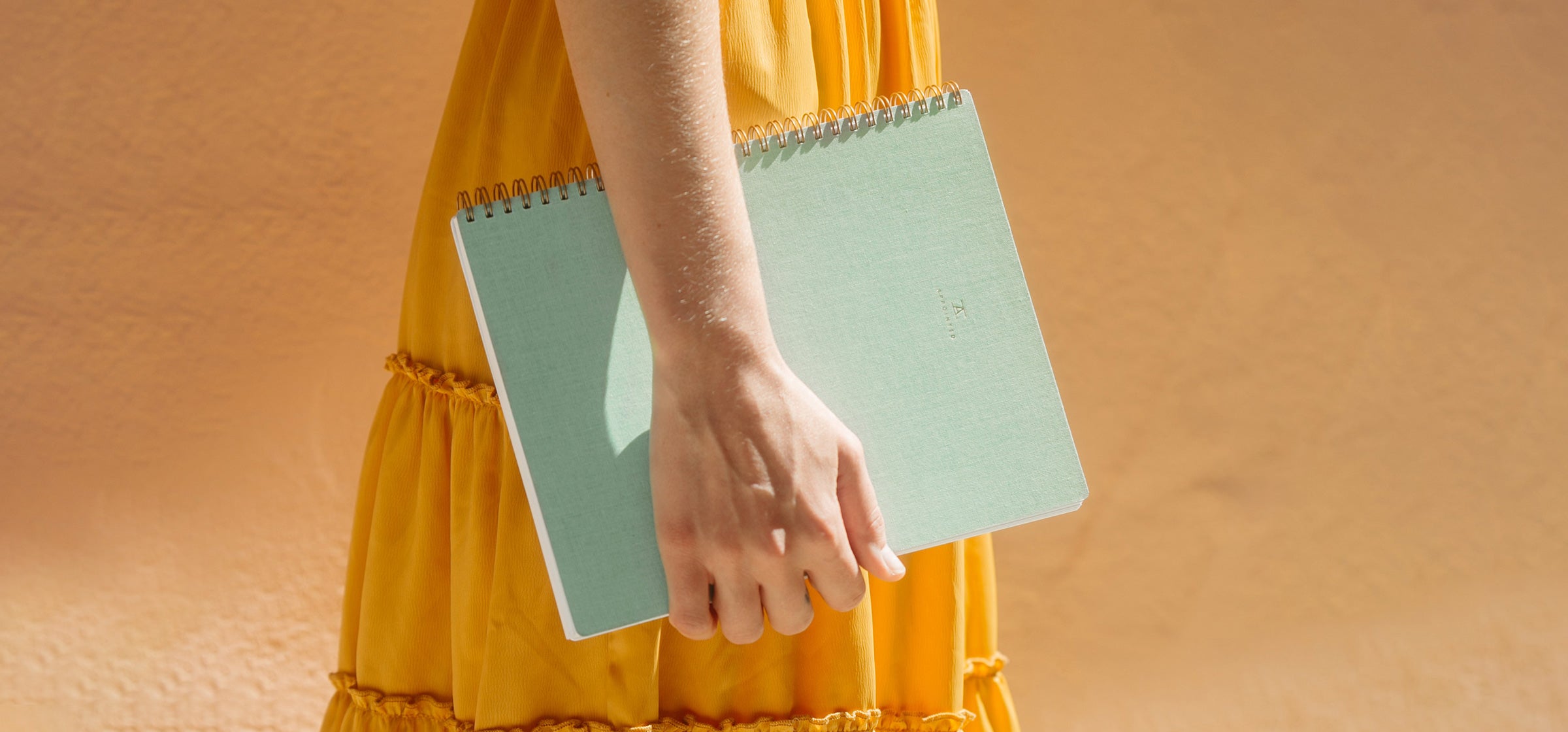 A woman in a marigold dress stands sideways against a peach-orange wall holding the Mineral Green Notebook in her right hand.