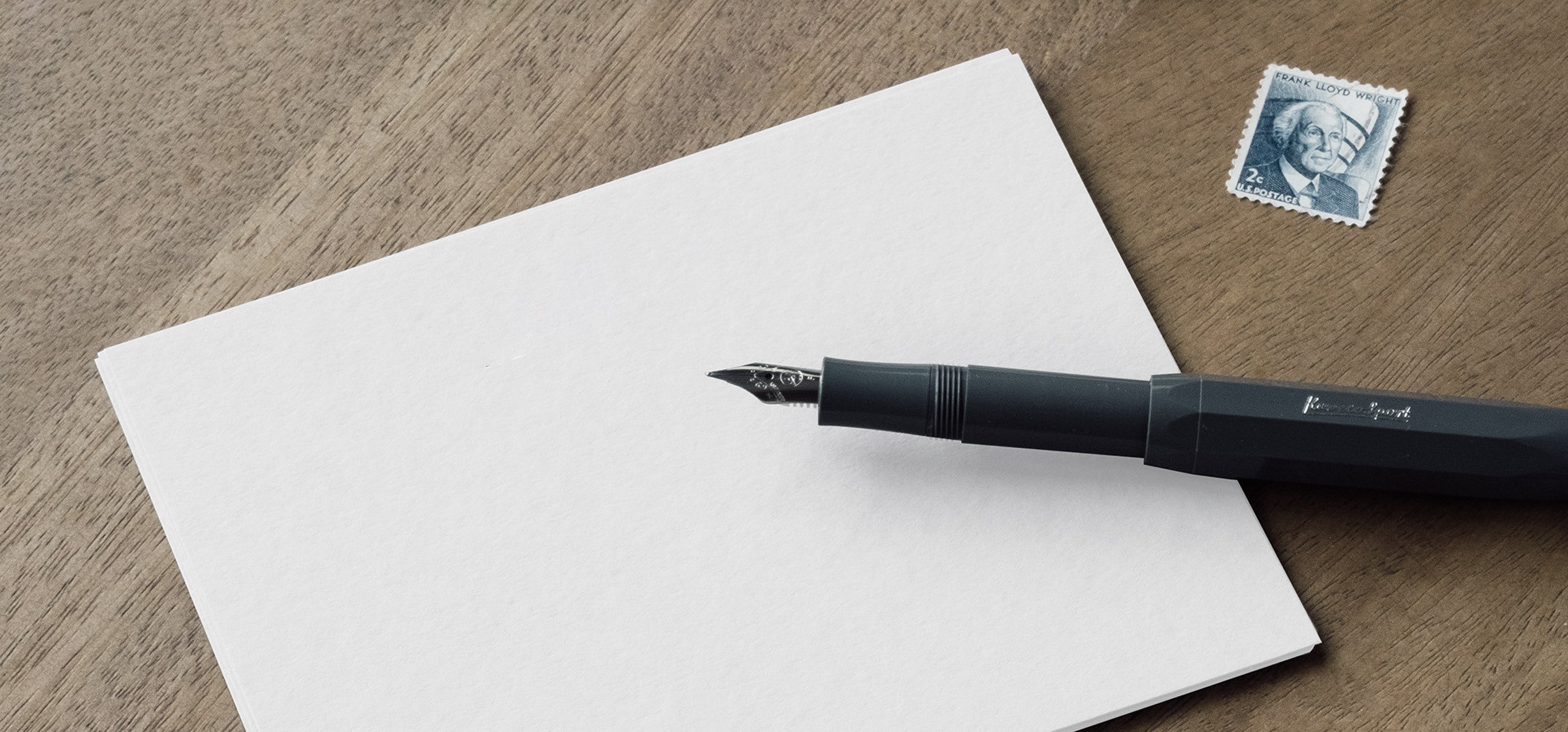 A Kaweco skyline sport fountain pen sits uncapped and prepared to write on a white card. A postage stamp sits to the side all on a wooden desktop.