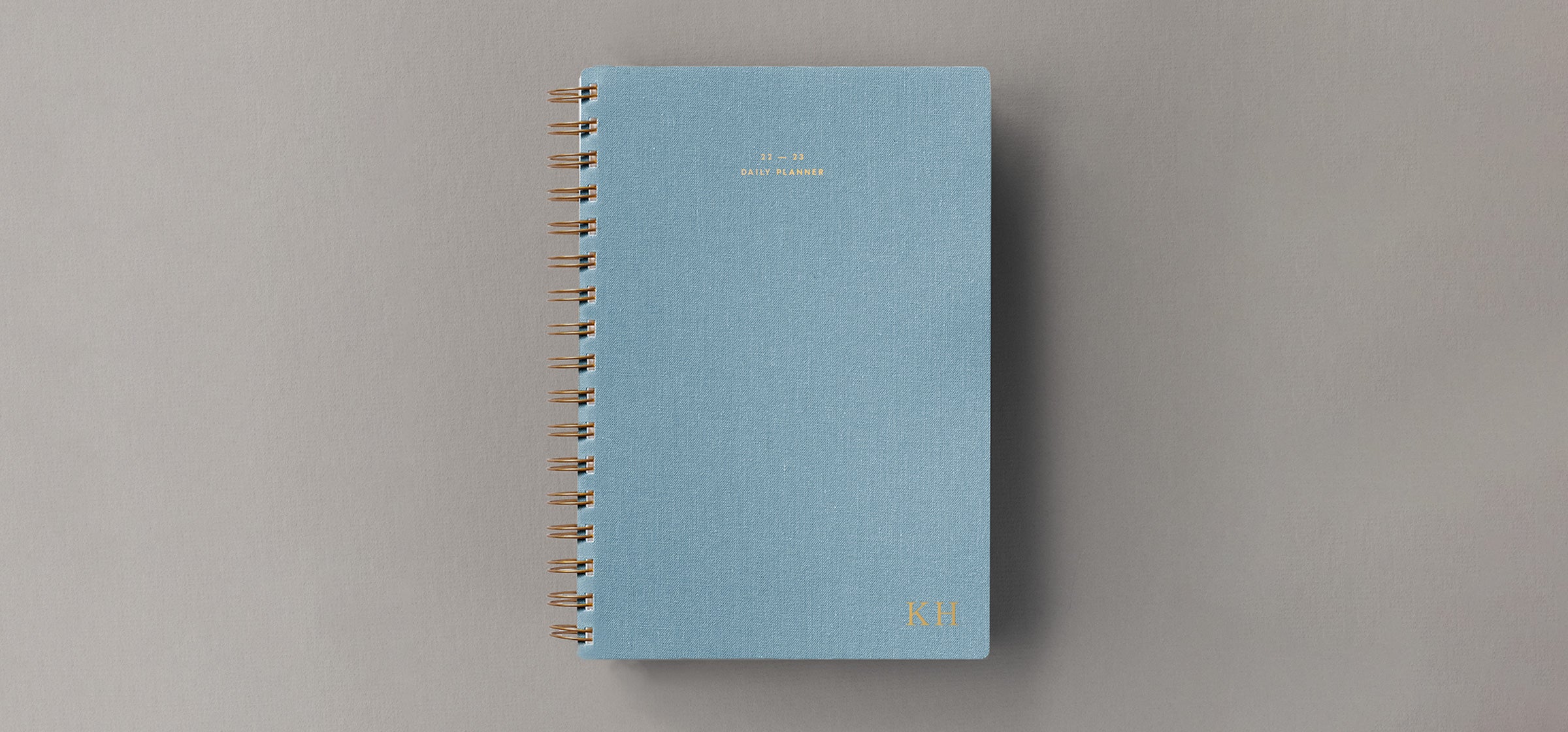 The 22-23 Wire Daily Planner in Chambray Blue bookcloth with brass binding and gold foil details sits against a grey-beige background