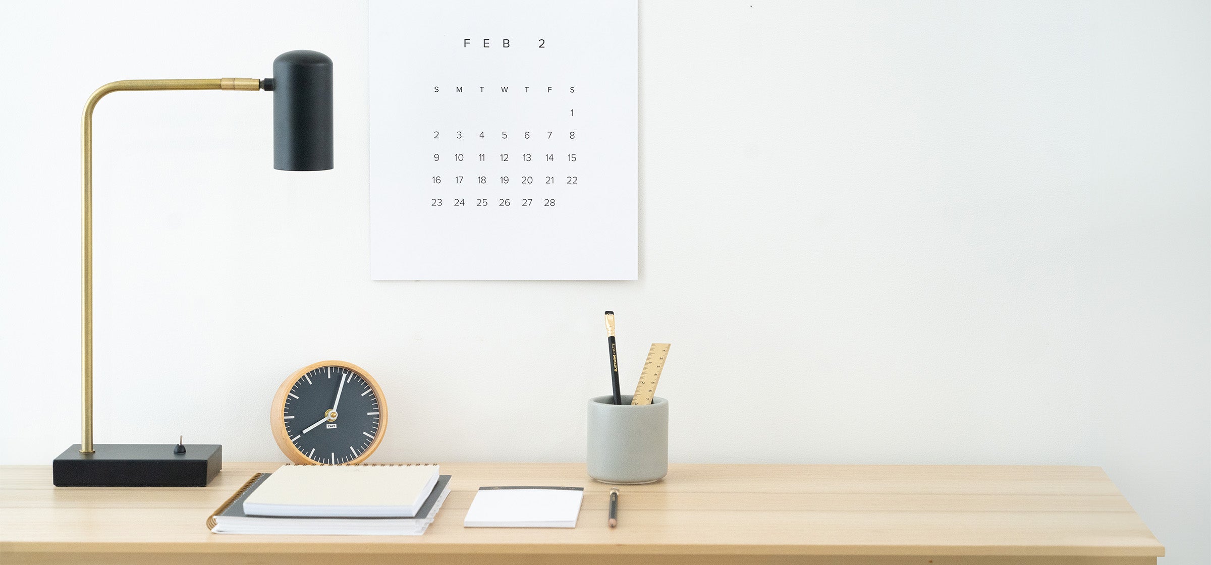 A calm desk scene shows a light wooden desk. Above the desk a minimal wall calendar hangs. There is a black and gold lamp, a clock, a pencil cup, and a stack of notebooks and planners on top of the desk.