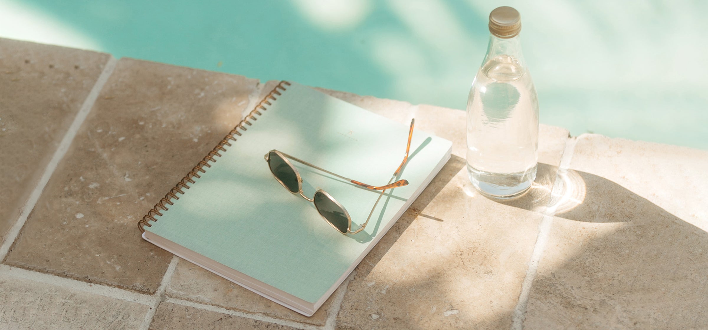 The Mineral Green Notebook sits on the edge of a swimming pool, dappled in shadow and light. A pair of sunglasses sits on top and a glass water bottle sits beside.