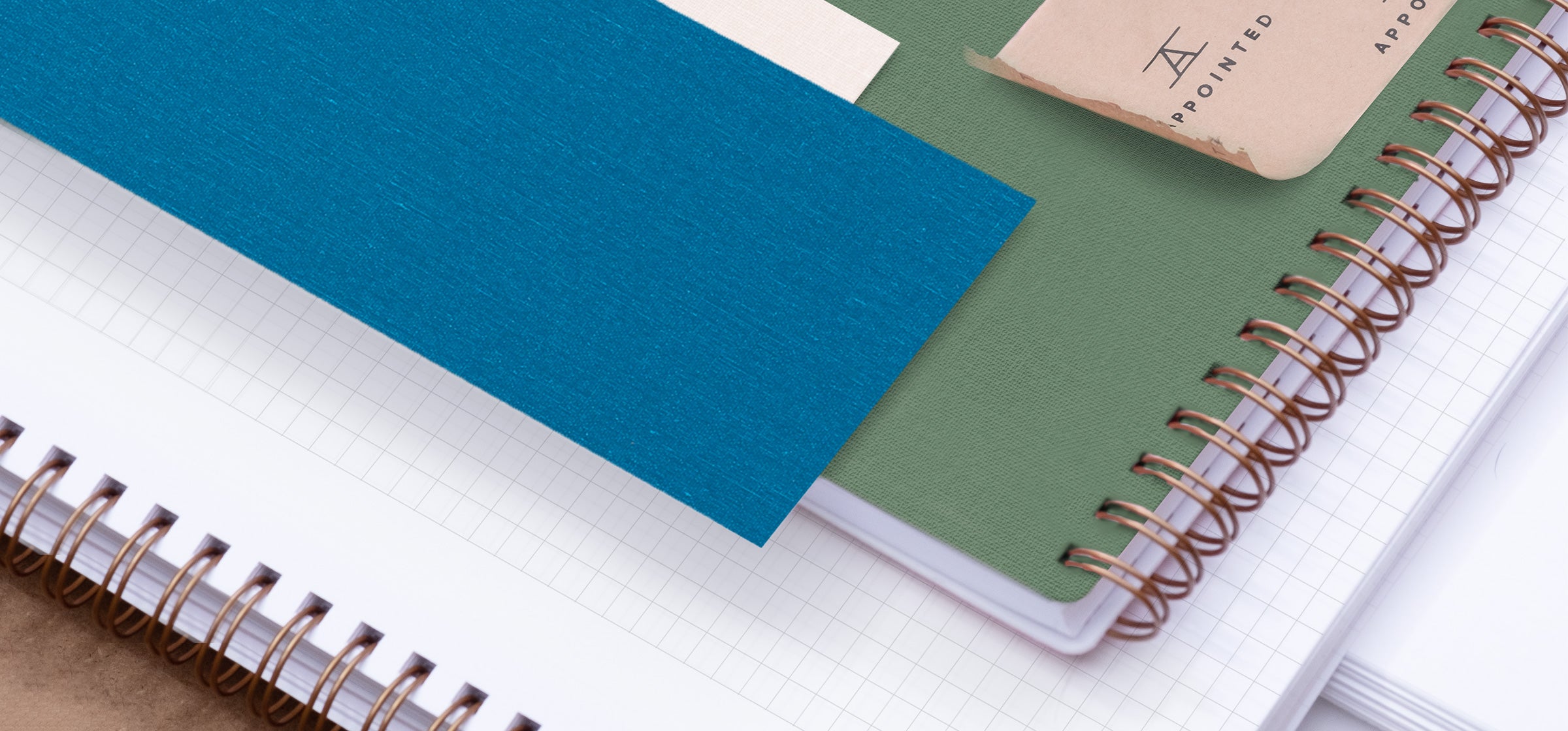 A detailed image shows an open notebook with brass wire-o binding stacked perpendicularly with a freen green workbook, bookcloth swatch, and appointed packaging tape
