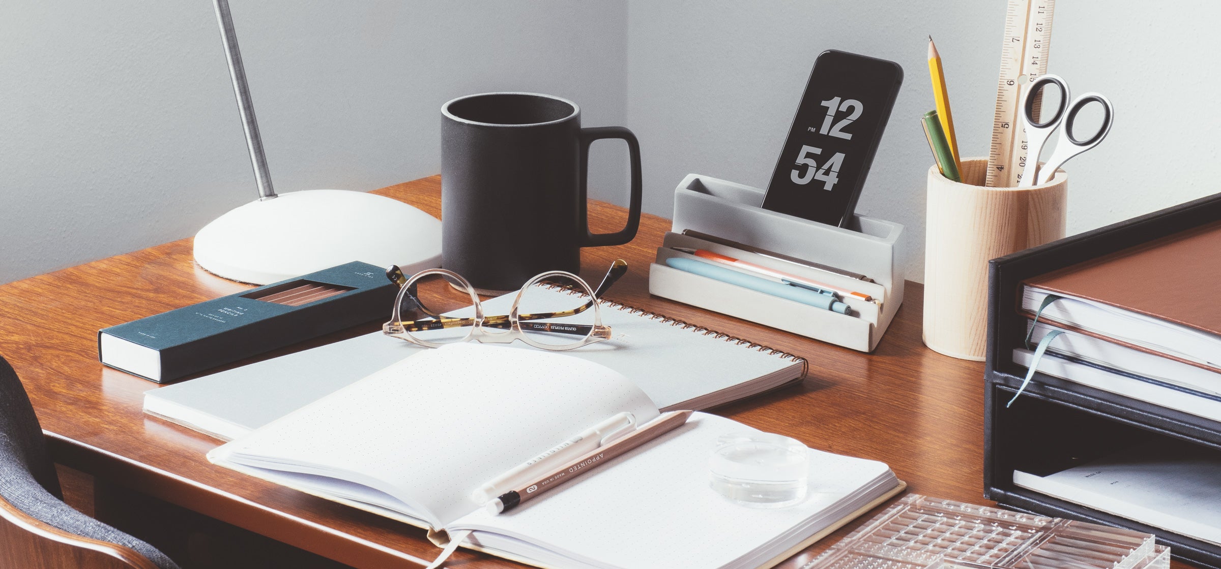 A desk scene shows an iphone, coffee cup, writing tools, and various stationery items like desk pads and notebooks strewn across a wooden desk