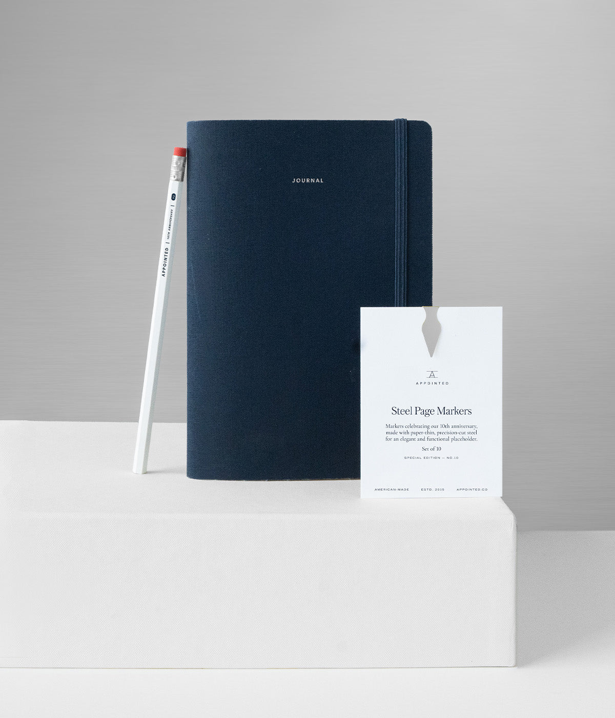 The Appointed 10th Anniversary Collection sits on a white riser against a silver backdrop. Including a blue journal, white pencil, and silver page markers.